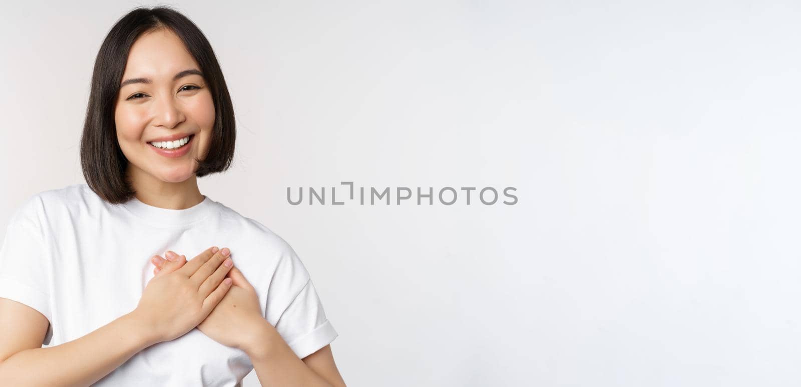 Close up of smiling korean woman holding hands on heart, care and love concept, feel affection, tenderness or heartwarming feeling, standing over white background by Benzoix