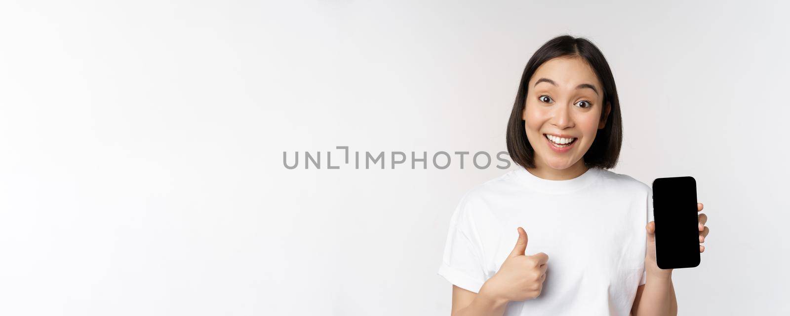 Enthusiastic young woman showing thumb up and mobile phone screen, standing in tshirt over white background. Copy space