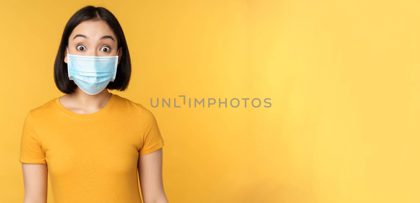 Portrait of korean girl in medical face mask looking surprised, amazed reaction to news, standing over yellow background.