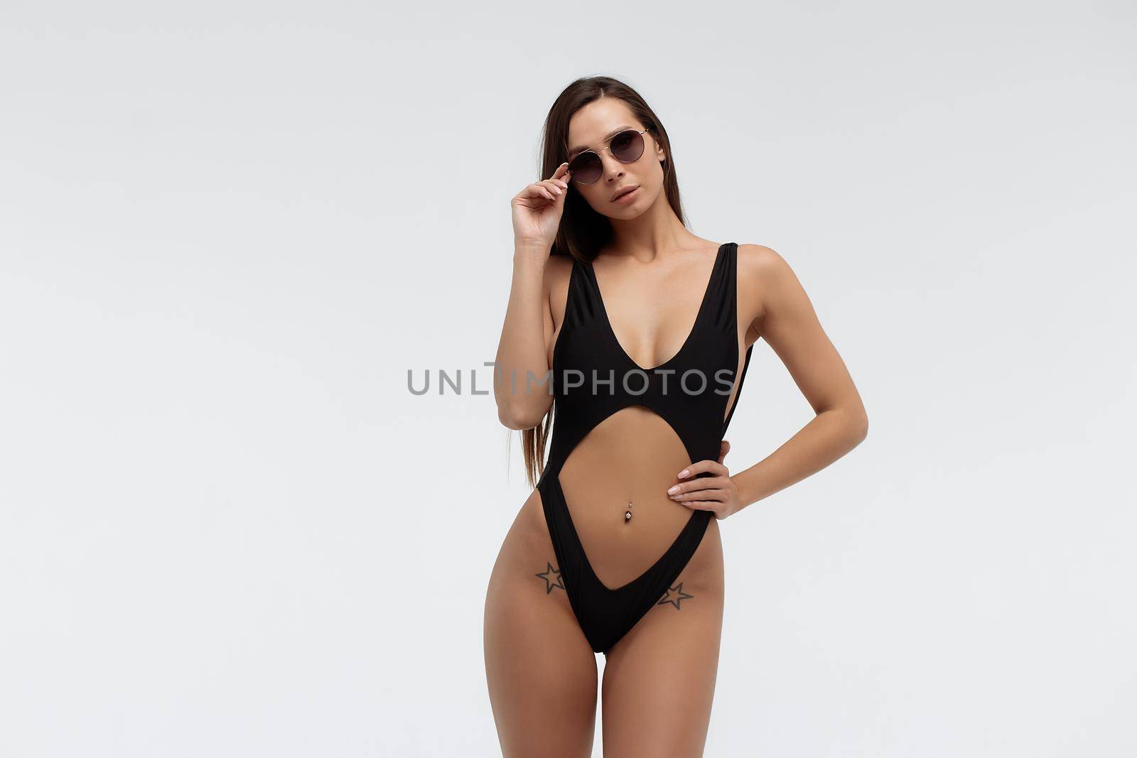 Side view of gorgeous young slim tanned brunette in trendy sunglasses and stylish sexy black swimwear looking at camera