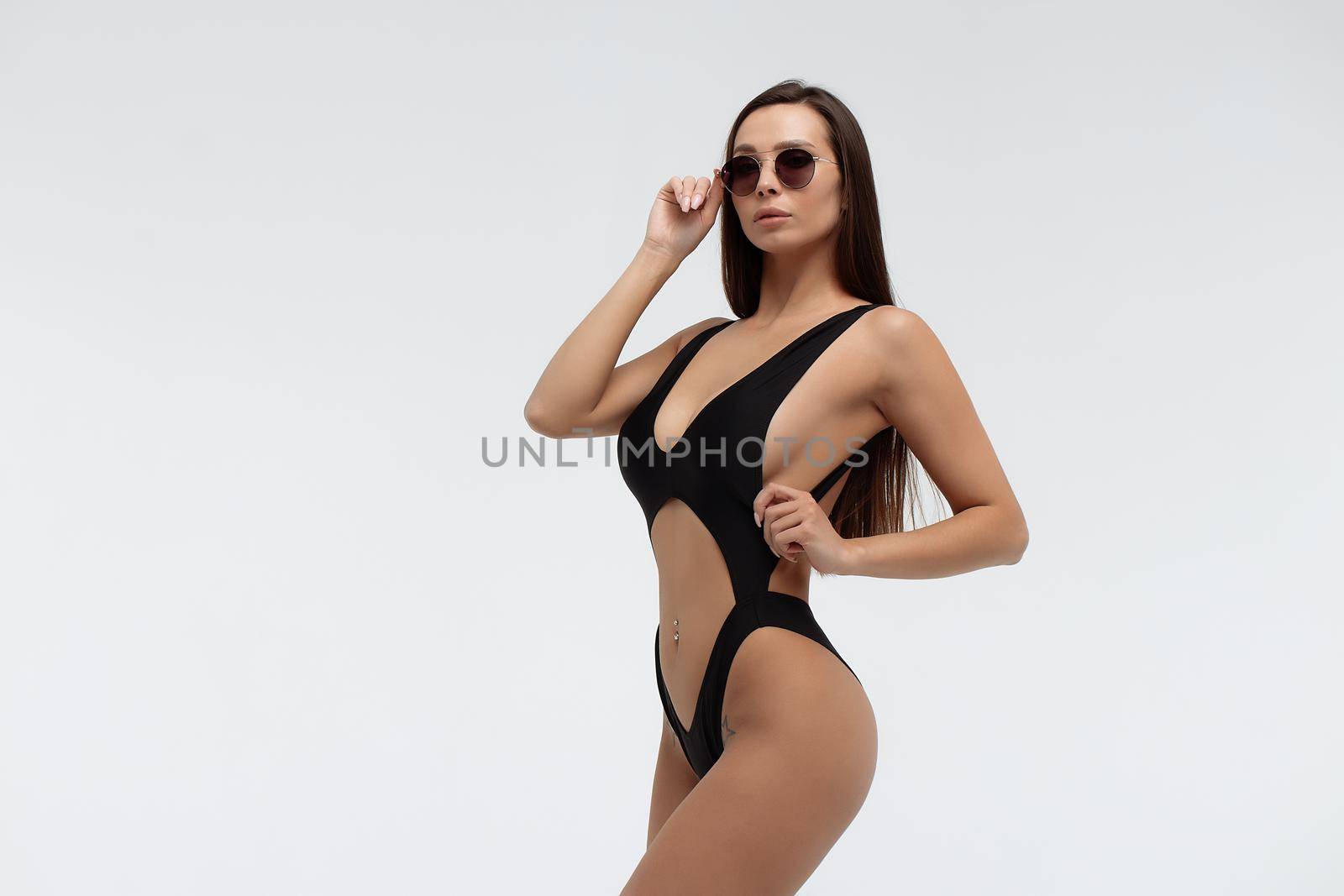 Side view of gorgeous young slim tanned brunette in trendy sunglasses and stylish sexy black swimwear looking at camera