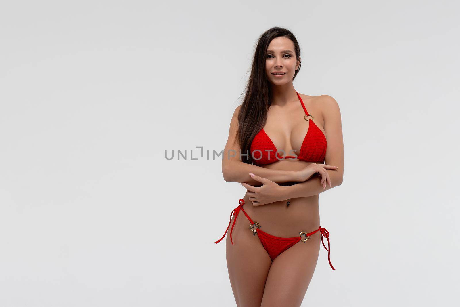 Seductive young long haired brunette with perfect tanned slim body wearing stylish red bikini looking at camera while standing against white background