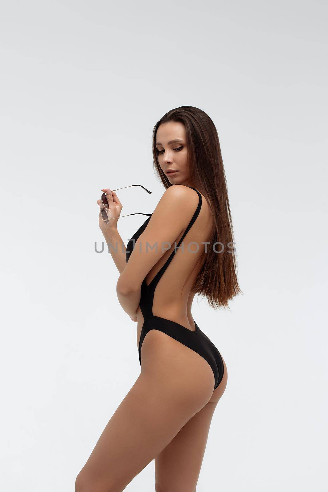 Side view of seductive coquettish young brunette with tanned slim body wearing sexy black swimsuit and holding sunglasses standing against white background