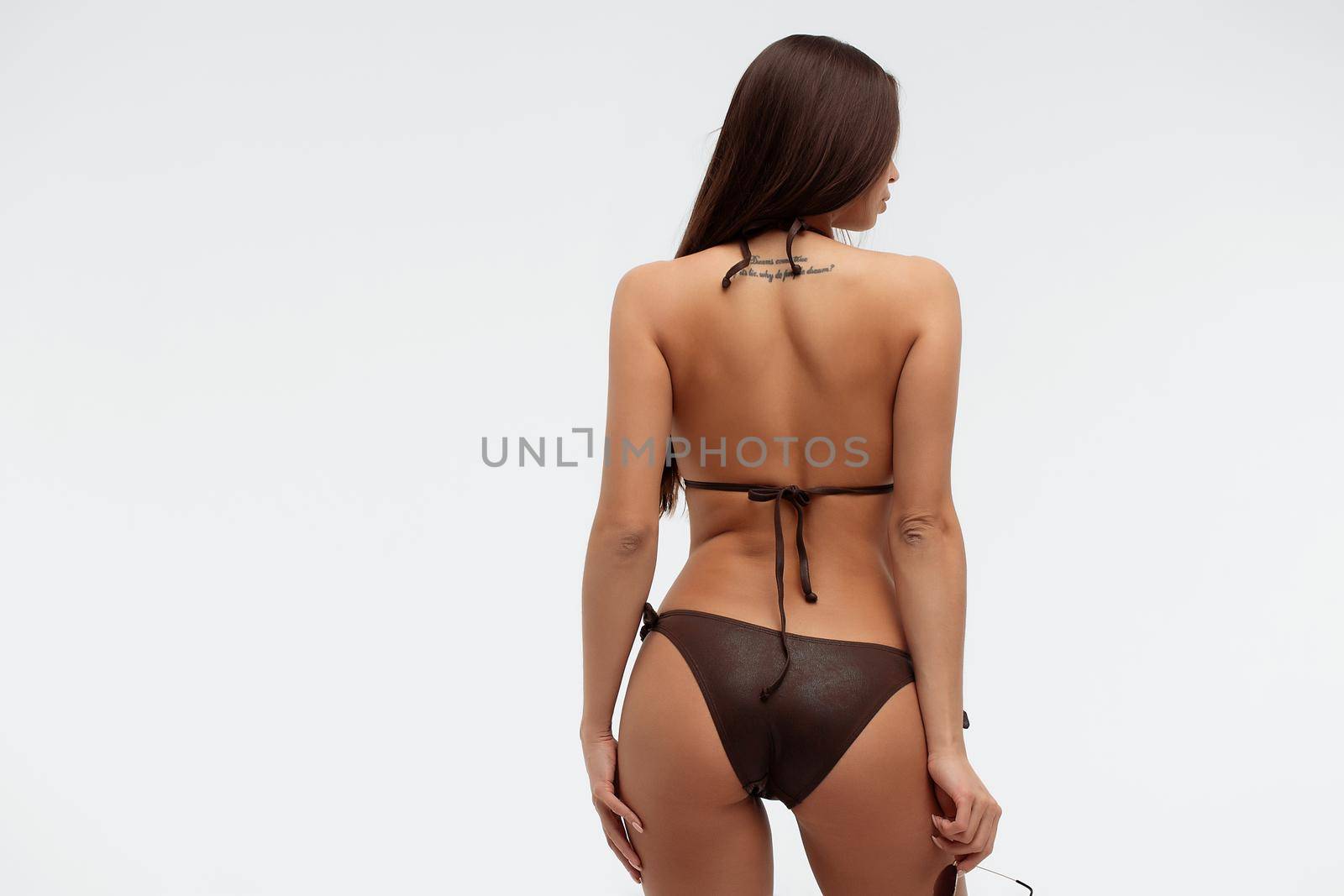 Side view of seductive coquettish young brunette with tanned slim body wearing sexy black swimsuit and holding sunglasses standing against white background