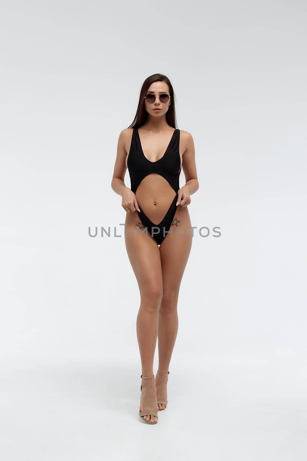 Attractive long haired young brunette model in sexy black bikini and trendy sunglasses touching hair while standing against white background