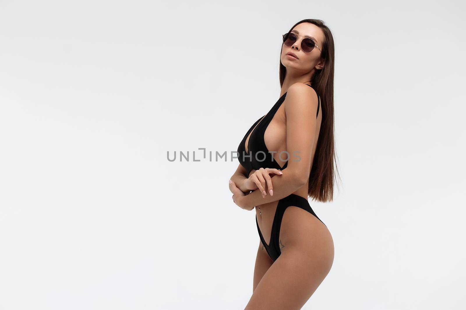 Side view of seductive coquettish young brunette with tanned slim body wearing sexy black swimsuit and holding sunglasses standing against white background