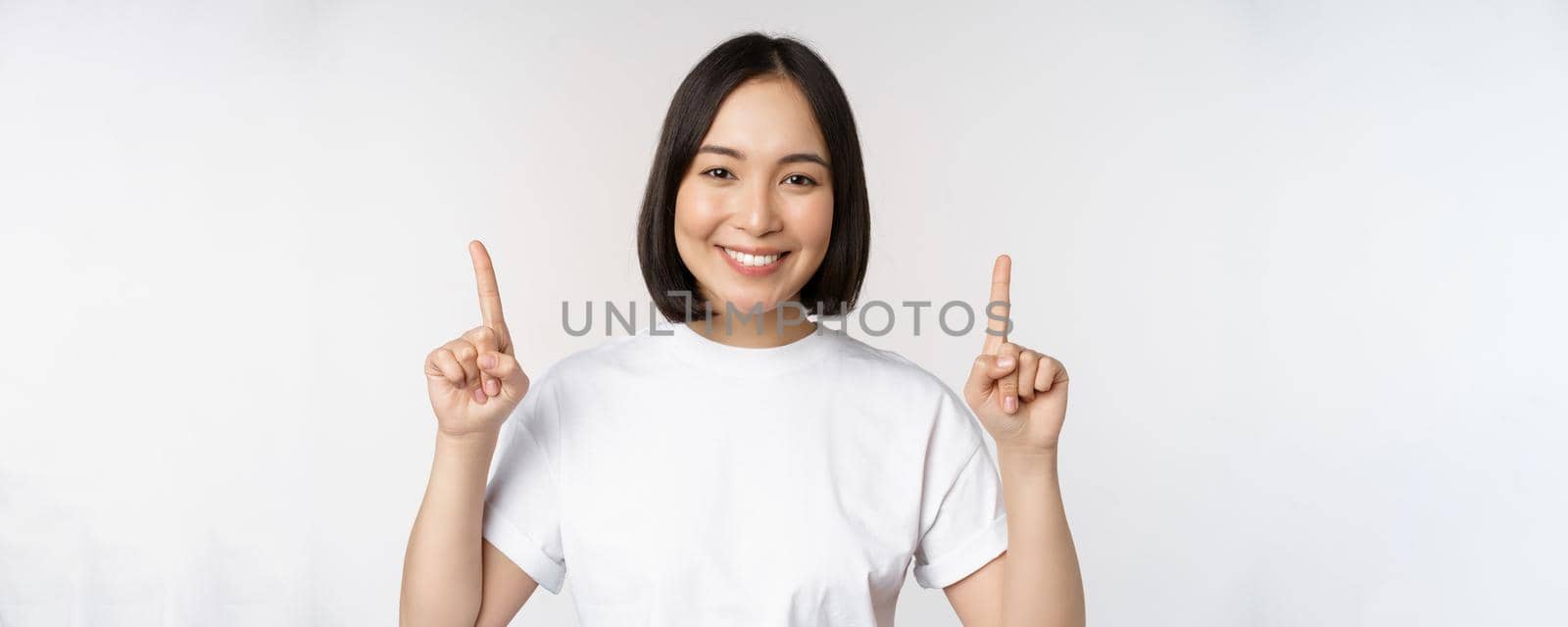 Portrait of enthusiastic young woman, asian girl smiling pointing fingers up, showing advertisement upwards, standing over white background by Benzoix