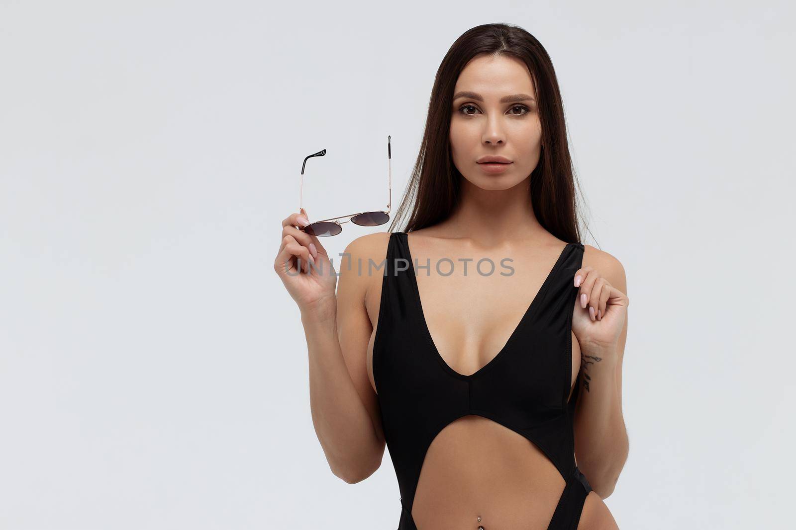 Attractive long haired young brunette model in sexy black bikini and trendy sunglasses touching hair while standing against white background