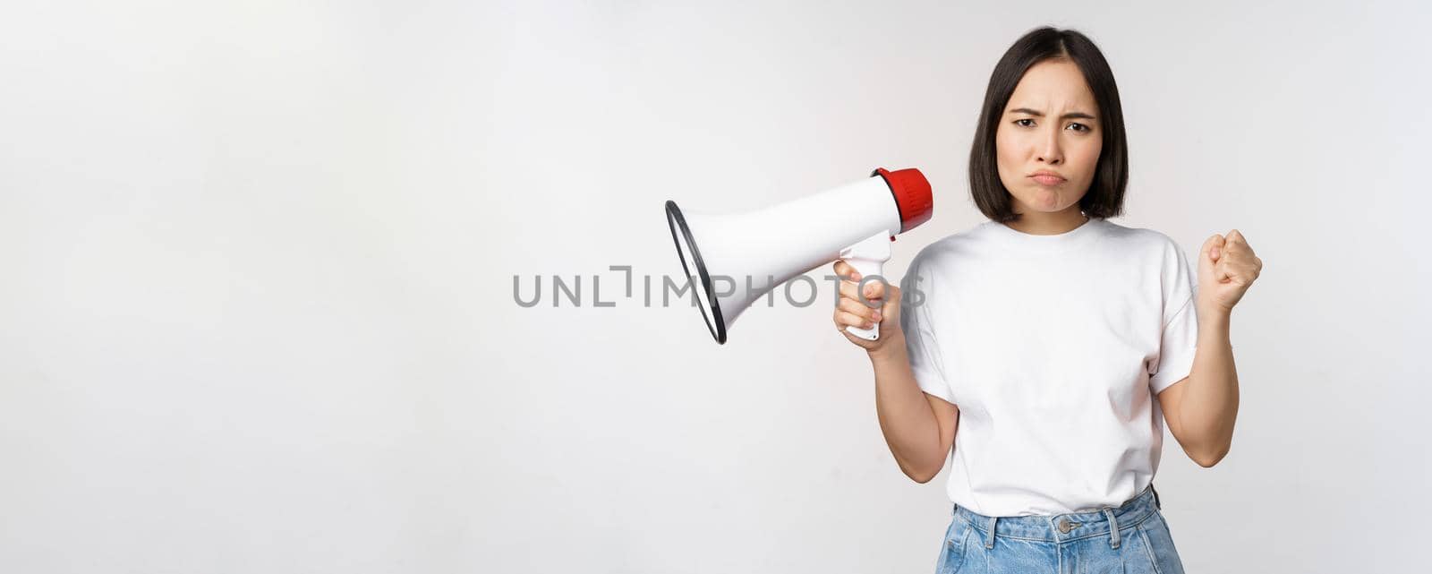 Angry asian girl activist, holding megaphone and looking furious, protesting, standing over white background. Copy space