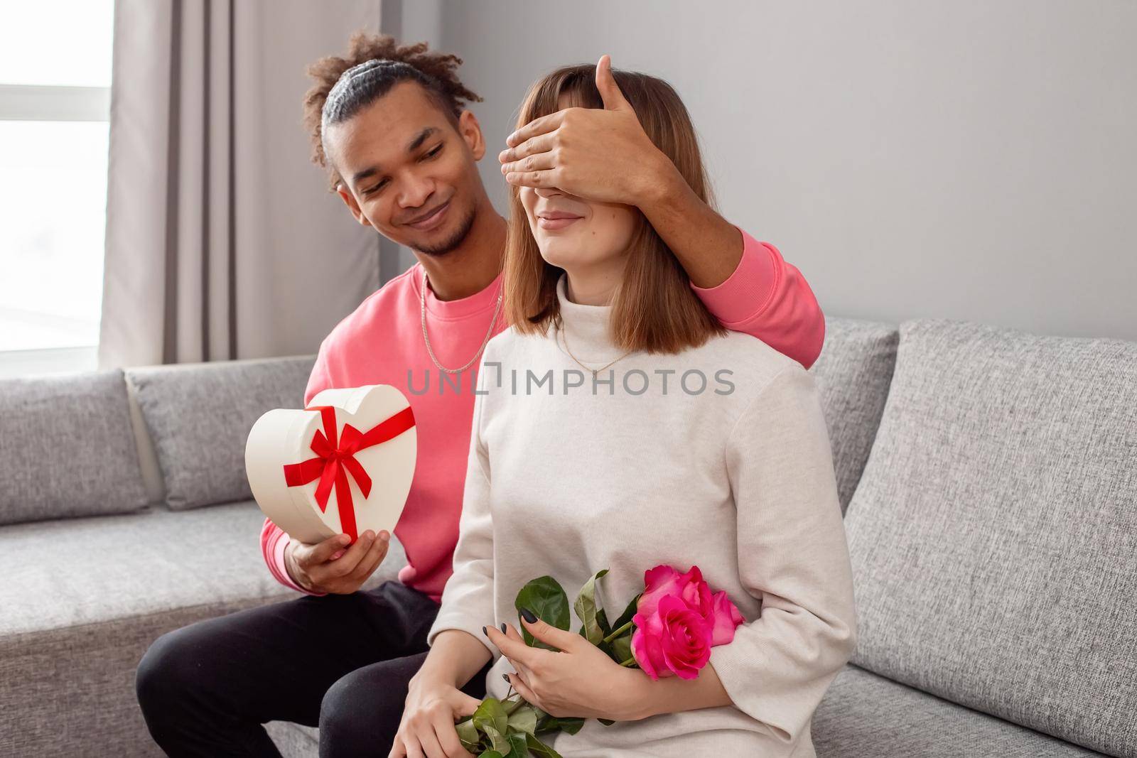A guy in a pink jumper and a girl in a white dress, on a gray couch . The guy covered the girls eyes with his hand to give a surprise - a white heart-shaped box with a bow and a bouquet of roses. copy space