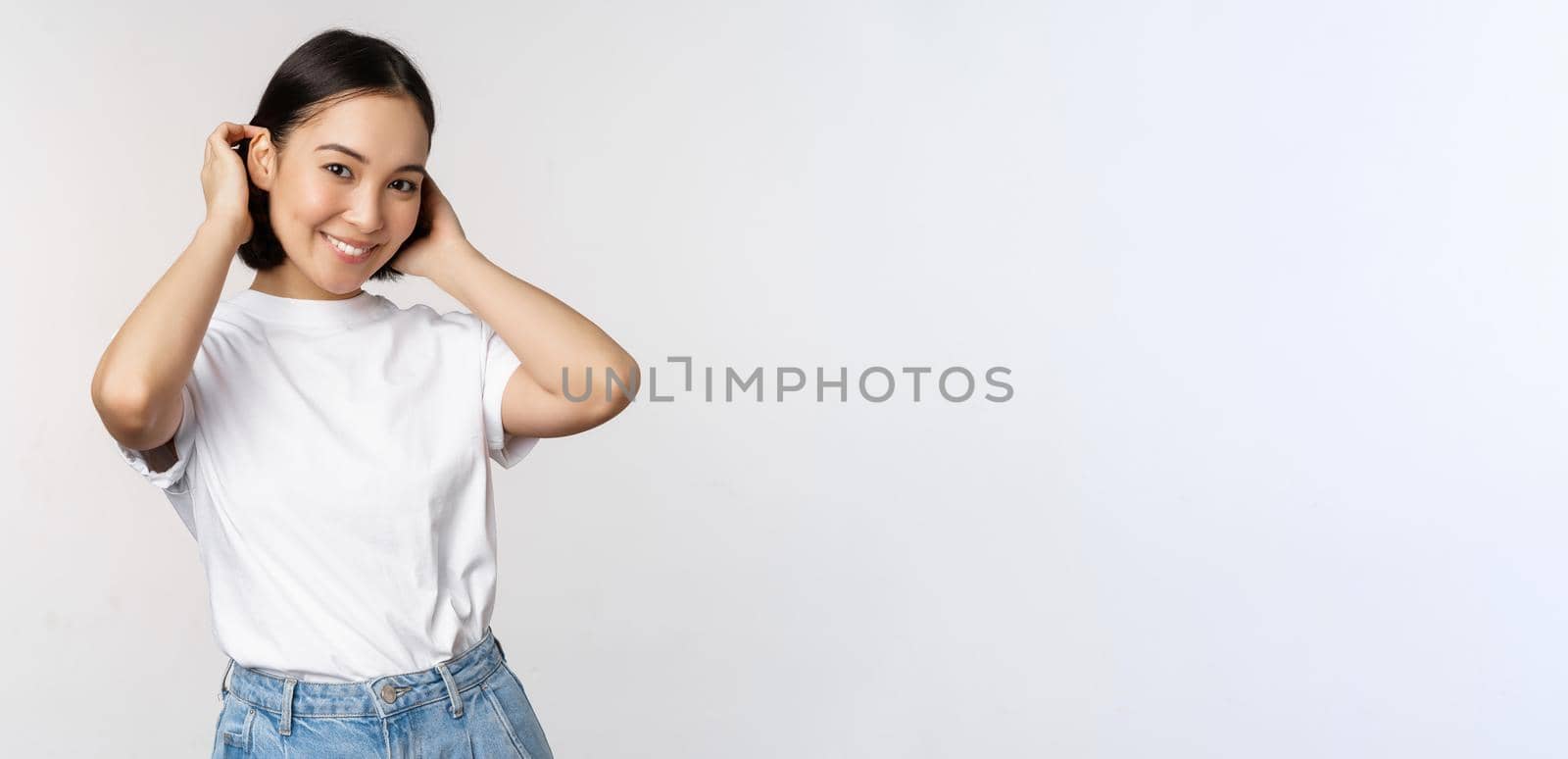 Portrait of cute, beautiful asian woman touching her new short haircut, showing hairstyle, smiling happy at camera, standing over white background by Benzoix