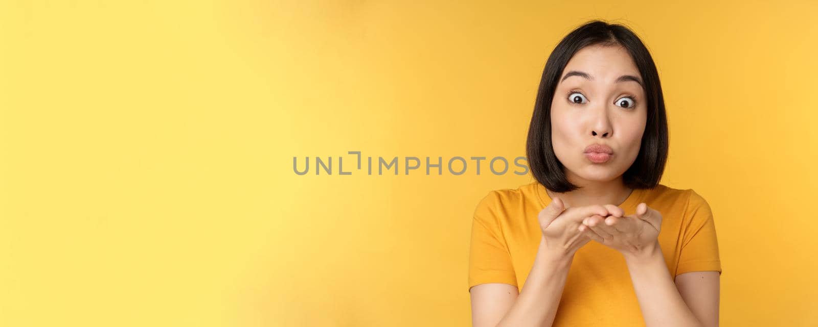 Cute asian girl sending air kiss, blowing mwah with puckered lips, standing over yellow background. Copy space