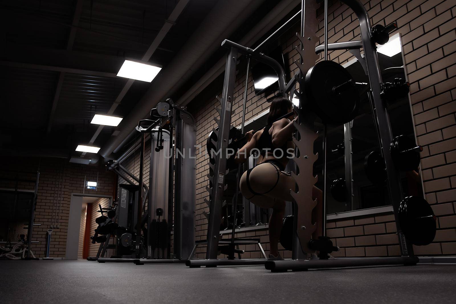 Low angle back view of slim athletic woman in sportswear with perfect sexy buttocks doing squats with heavy barbell during workout in gym