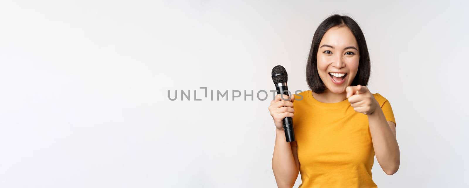Enthusiastic asian girl with microphone, pointing finger at camera, suggesting you to sing, standing over white background by Benzoix
