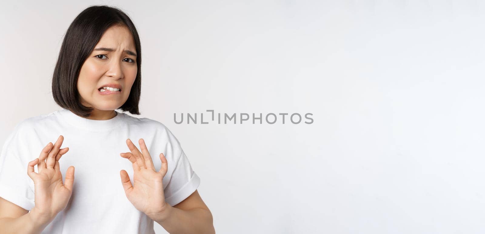 Disgusted asian woman rejecting smth, grimacing from dislike and aversion, stare with cringe, refusing offer, standing over white background by Benzoix