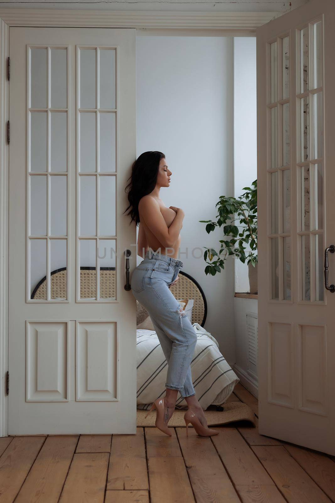Full body of barefoot young slim female in trendy ripped jeans covering naked breast with hand while leaving bedroom at home