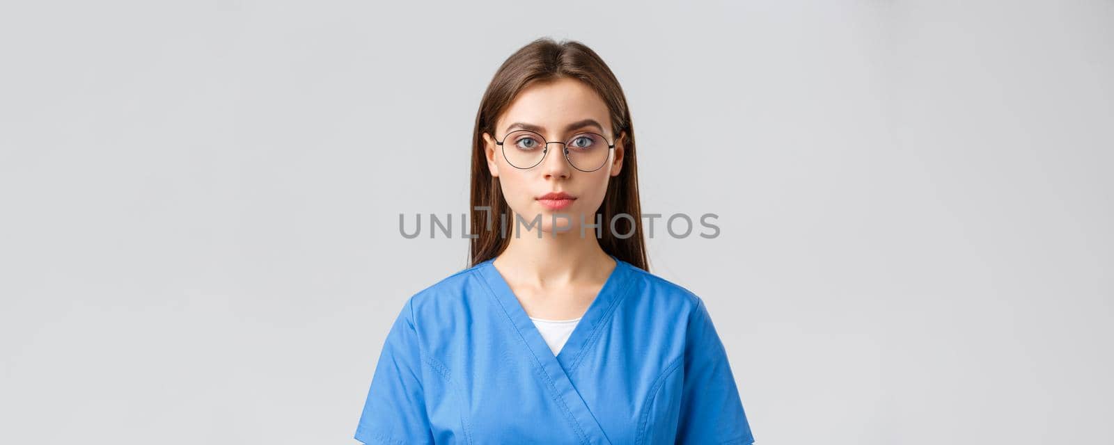 Healthcare workers, medicine, insurance and covid-19 pandemic concept. Young female intern, nurse or doctor in scrubs and glasses, looking at camera with serious expression, grey background by Benzoix