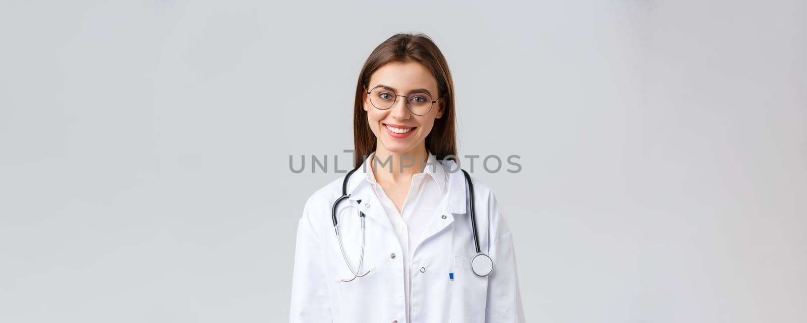 Healthcare workers, medicine, insurance and covid-19 pandemic concept. Smiling attractive female doctor, physician in white scrubs with stethoscope and glasses look upbeat at camera by Benzoix