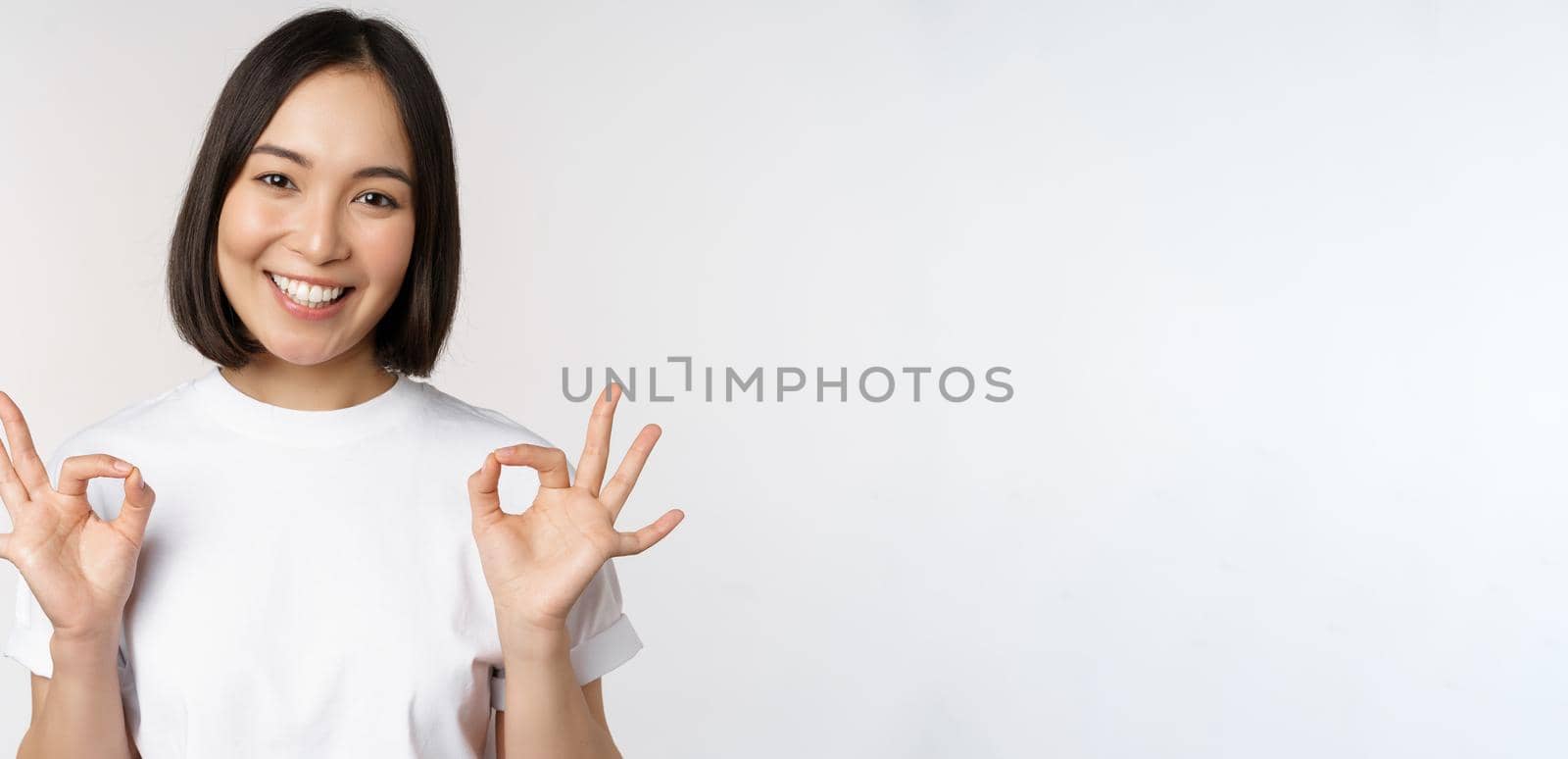 Very well, excellent. Smiling asian woman showing okay sign, approval, ok gesture, looking satisfied, recommending smth, standing over white background by Benzoix