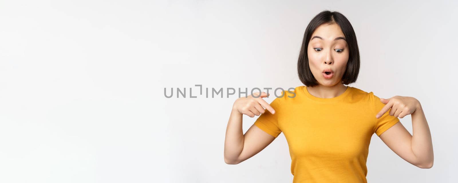 Happy asian girl pointing fingers down, looking at announcement, banner or advertisment, standing in yellow tshirt over white background.