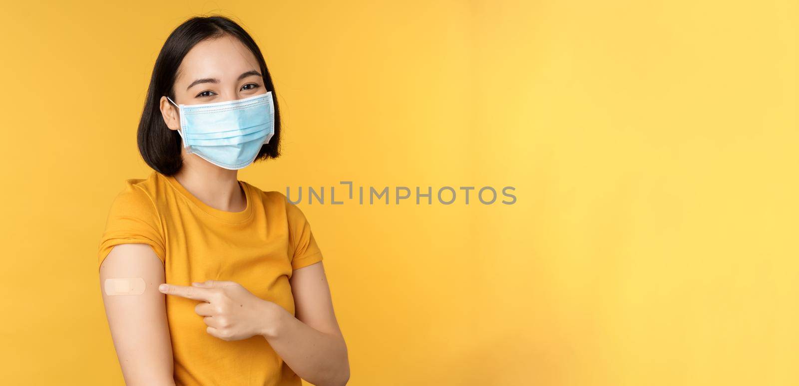 Vaccination and covid-19 pandemic concept. Smiling asian woman in medical face mask, showing her shoulder with band aid after vaccinating from coronavirus, yellow background.