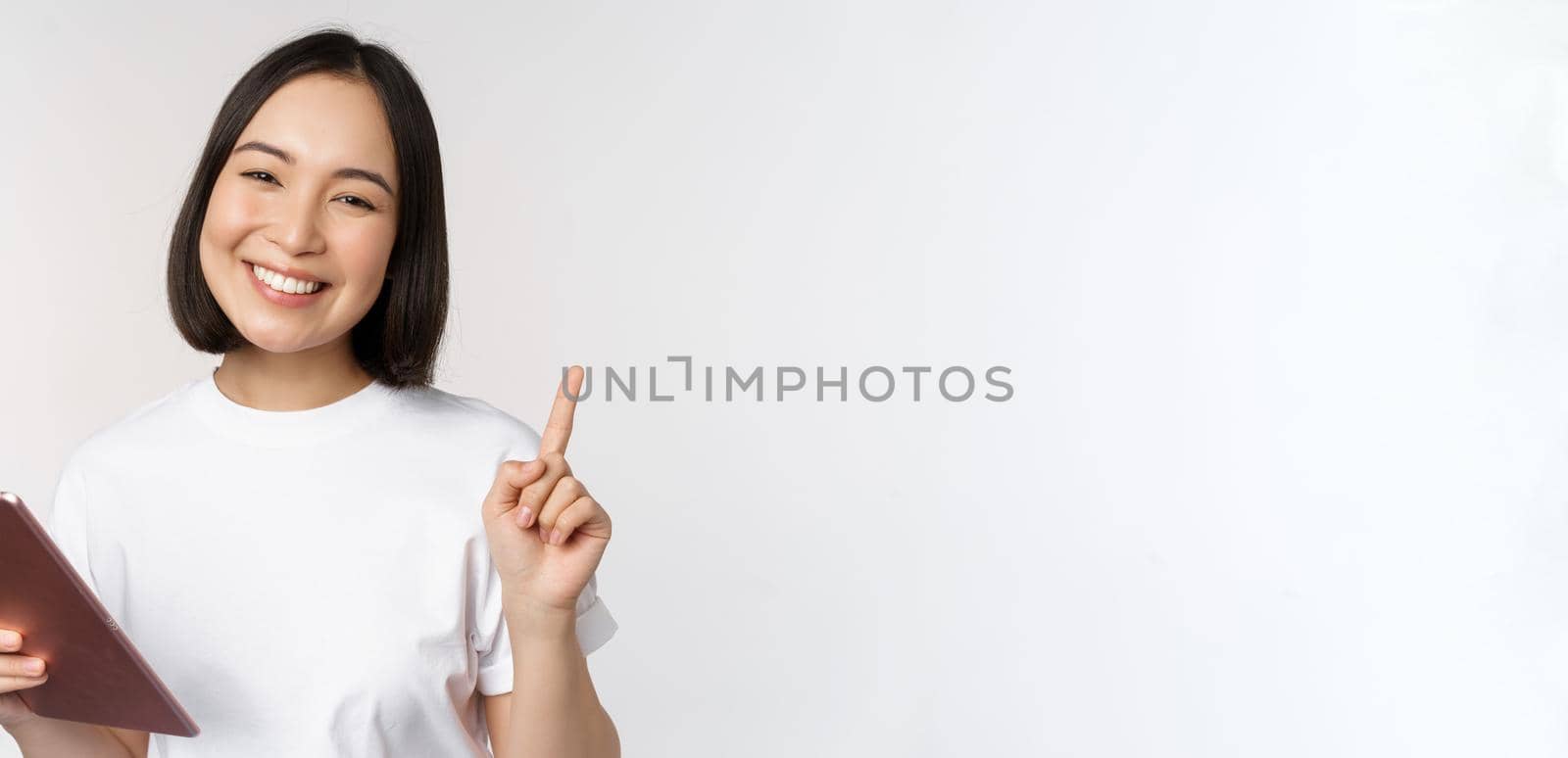 Enthusiastic asian woman with tablet, raising finger and looking amazed, pointing up, standing over white background by Benzoix