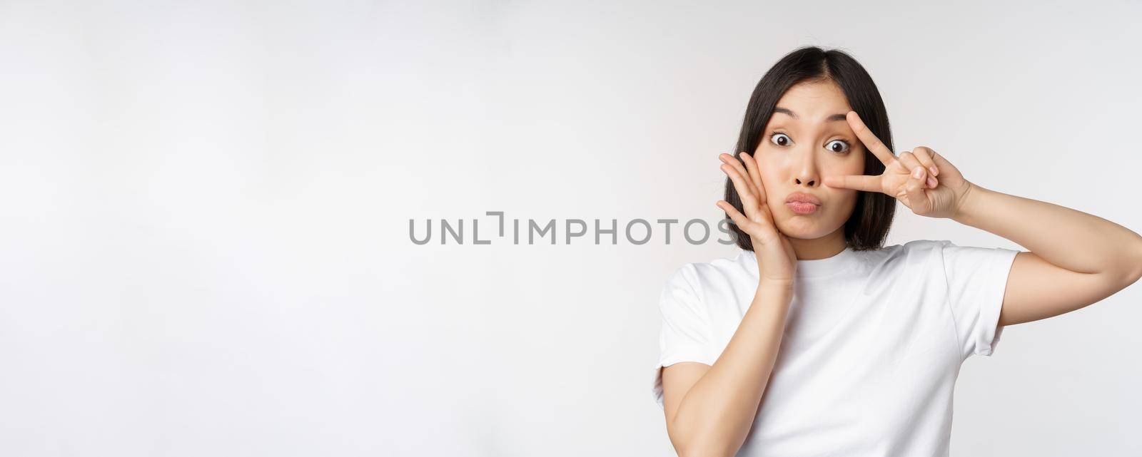 Cute asian girl posing with kawaii v-sign, peace gesture near face, standing in tshirt over white background by Benzoix