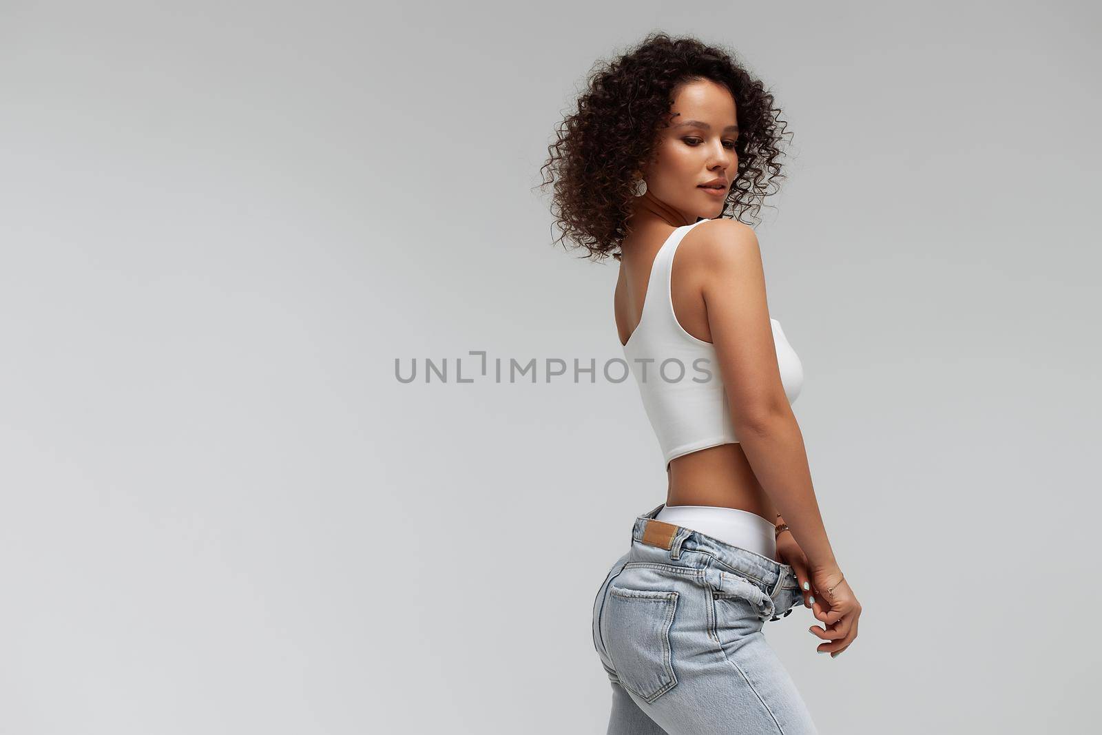 Full body of confident young barefoot female model with curly hairstyle wearing trendy denim jacket and jeans