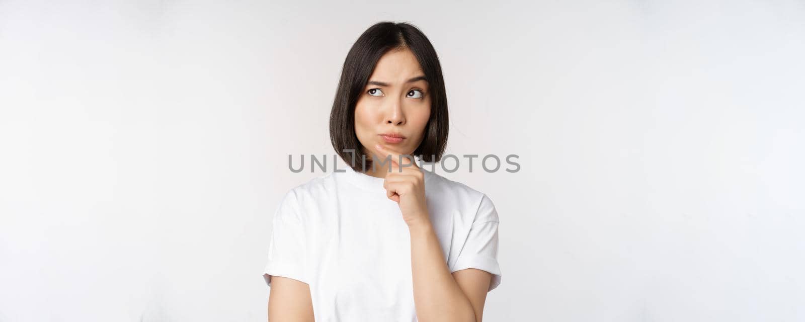 Thoughtful asian woman looking aside, pondering, making assumption, thinking or choosing smth, standing over white background.