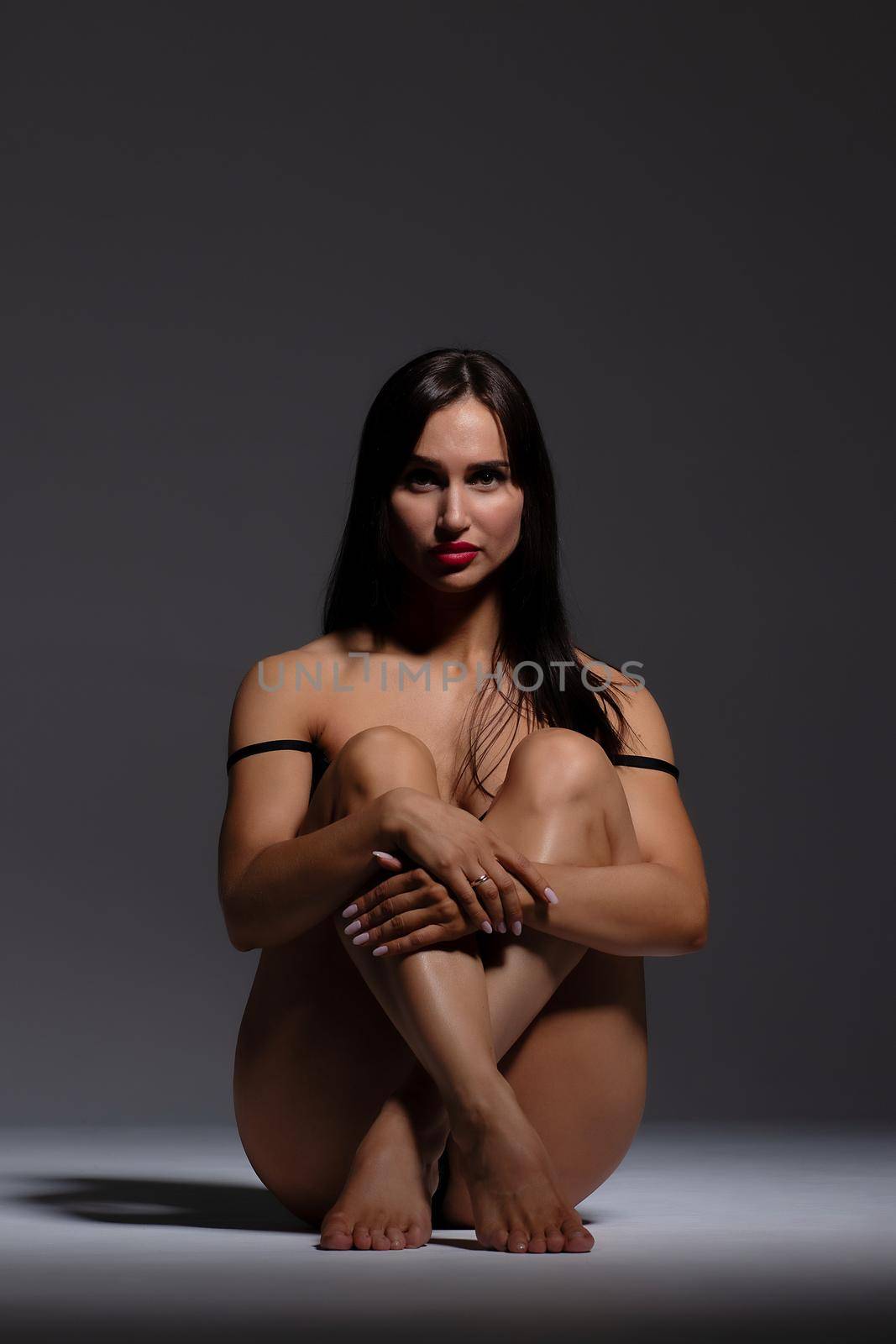 Side view full body attractive fit brunette wearing black underwear and high heels sitting on haunches and looking at camera in dark studio