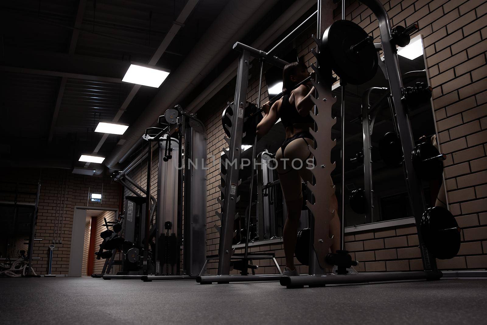 Low angle back view of slim athletic woman in sportswear with perfect sexy buttocks doing squats with heavy barbell during workout in gym