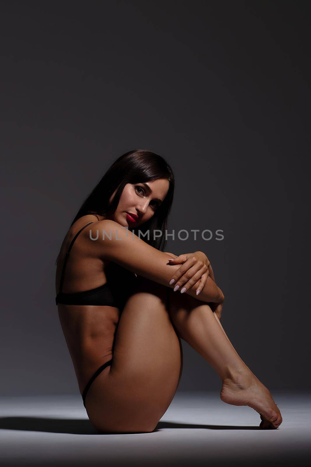 Side view full body attractive fit brunette wearing black underwear and high heels sitting on haunches and looking at camera in dark studio