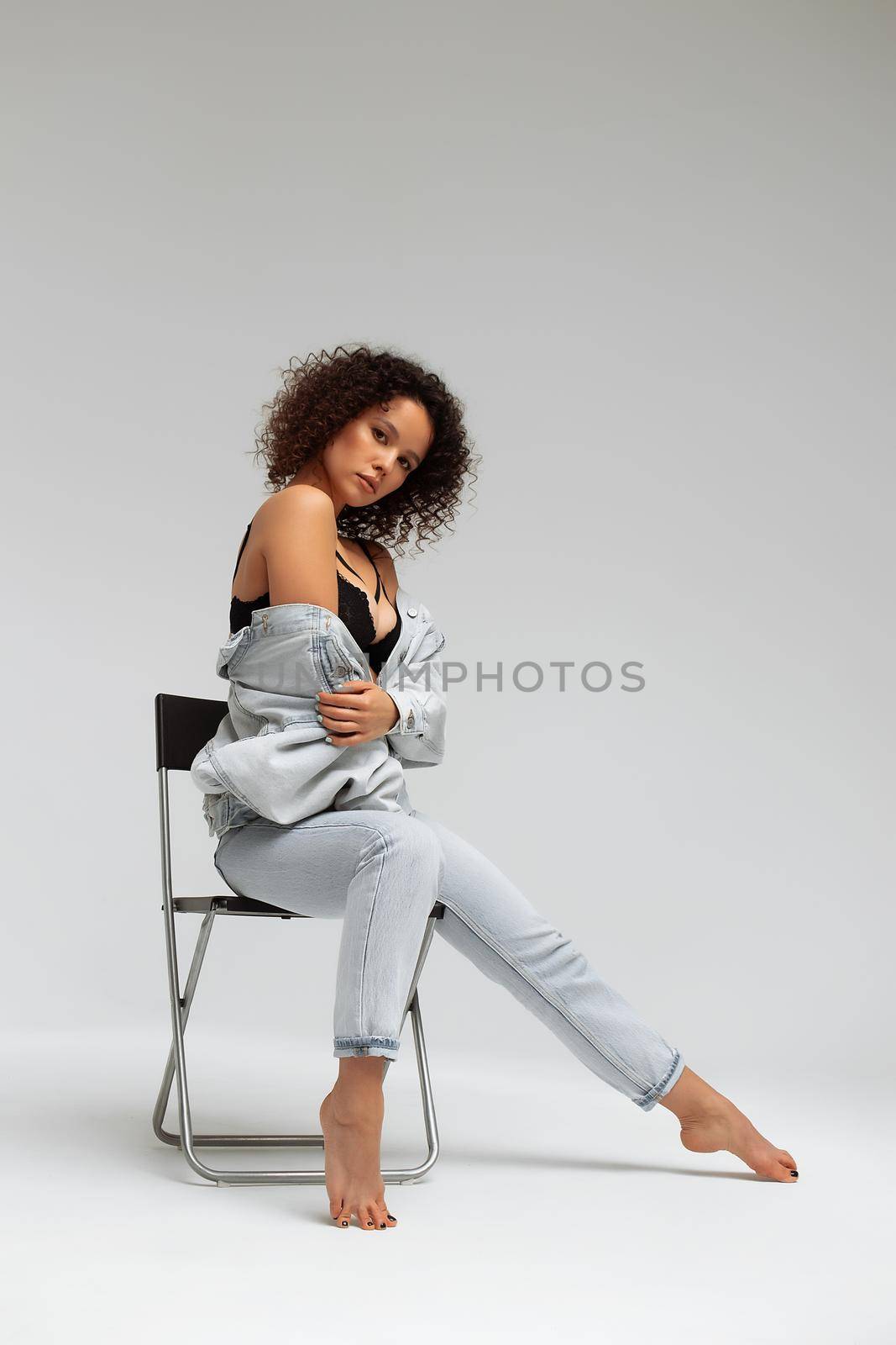 Full body of confident young barefoot female model with curly hairstyle wearing trendy denim jacket and jeans sitting on chair in white studio