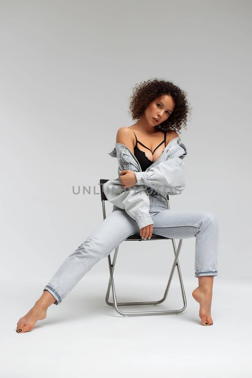 Full body of confident young barefoot female model with curly hairstyle wearing trendy denim jacket and jeans sitting on chair in white studio