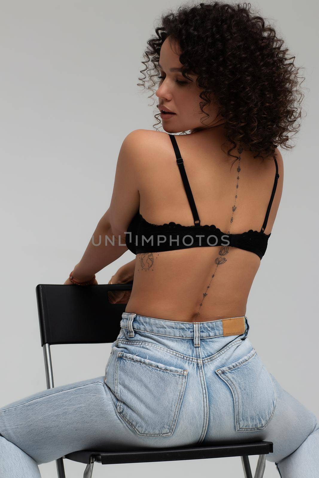 Full body of confident young barefoot female model with curly hairstyle wearing trendy denim jacket and jeans sitting on chair in white studio