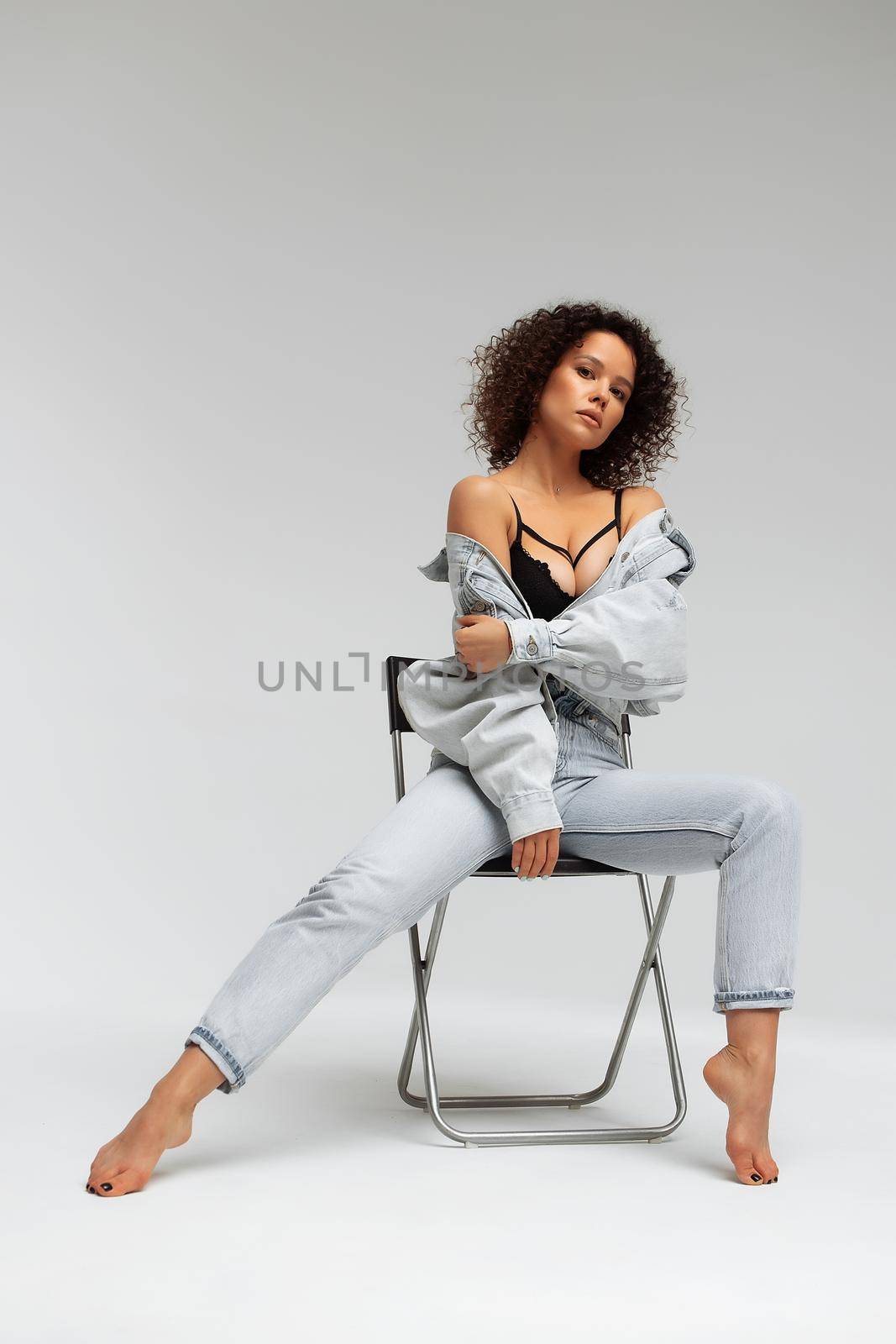 Full body of confident young barefoot female model with curly hairstyle wearing trendy denim jacket and jeans sitting on chair in white studio