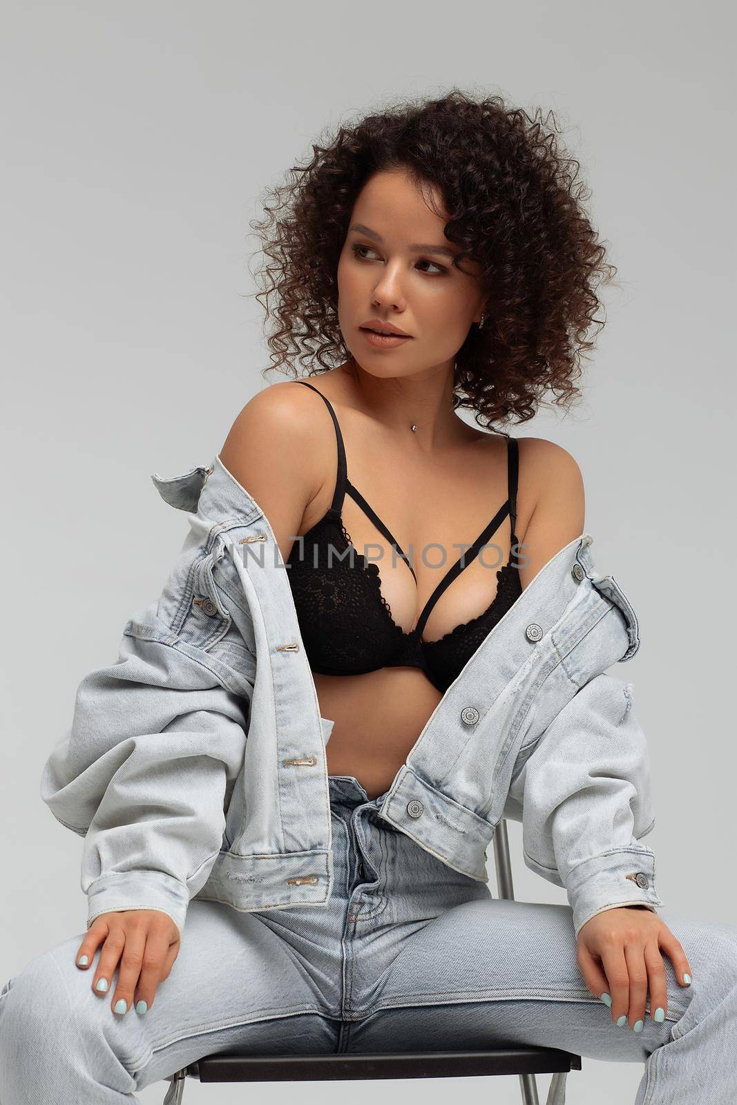 Full body of confident young barefoot female model with curly hairstyle wearing trendy denim jacket and jeans sitting on chair in white studio