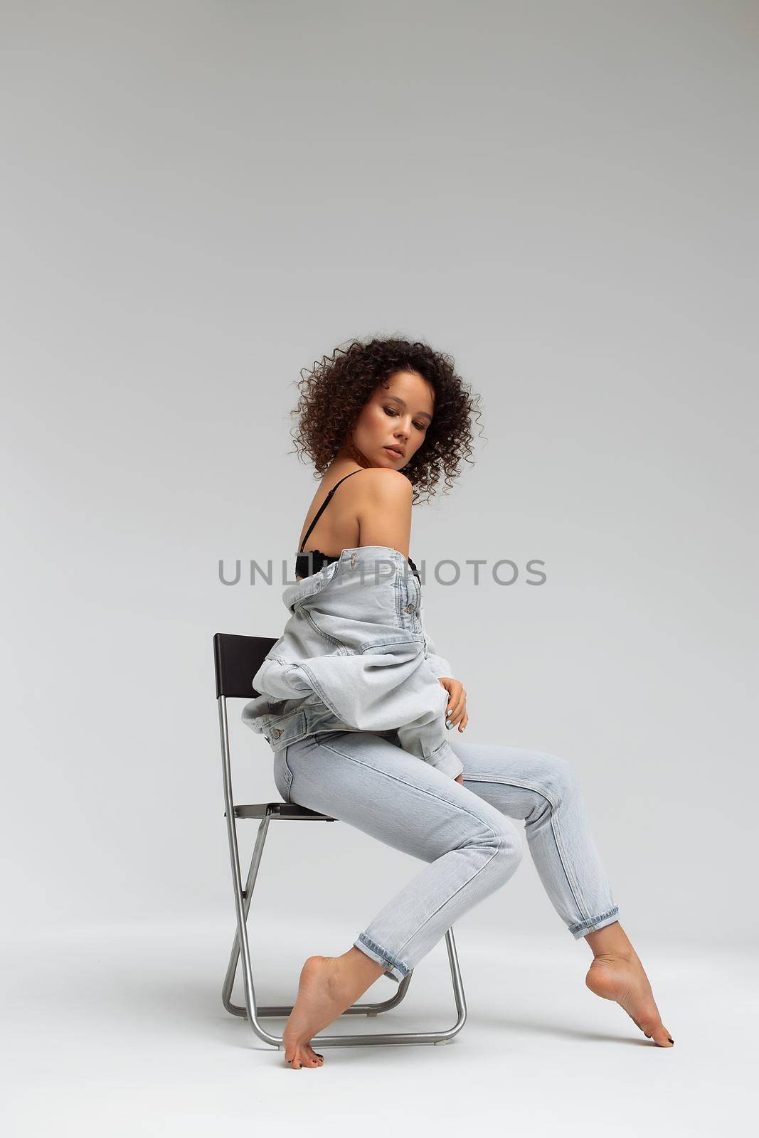 Full body of confident young barefoot female model with curly hairstyle wearing trendy denim jacket and jeans sitting on chair in white studio