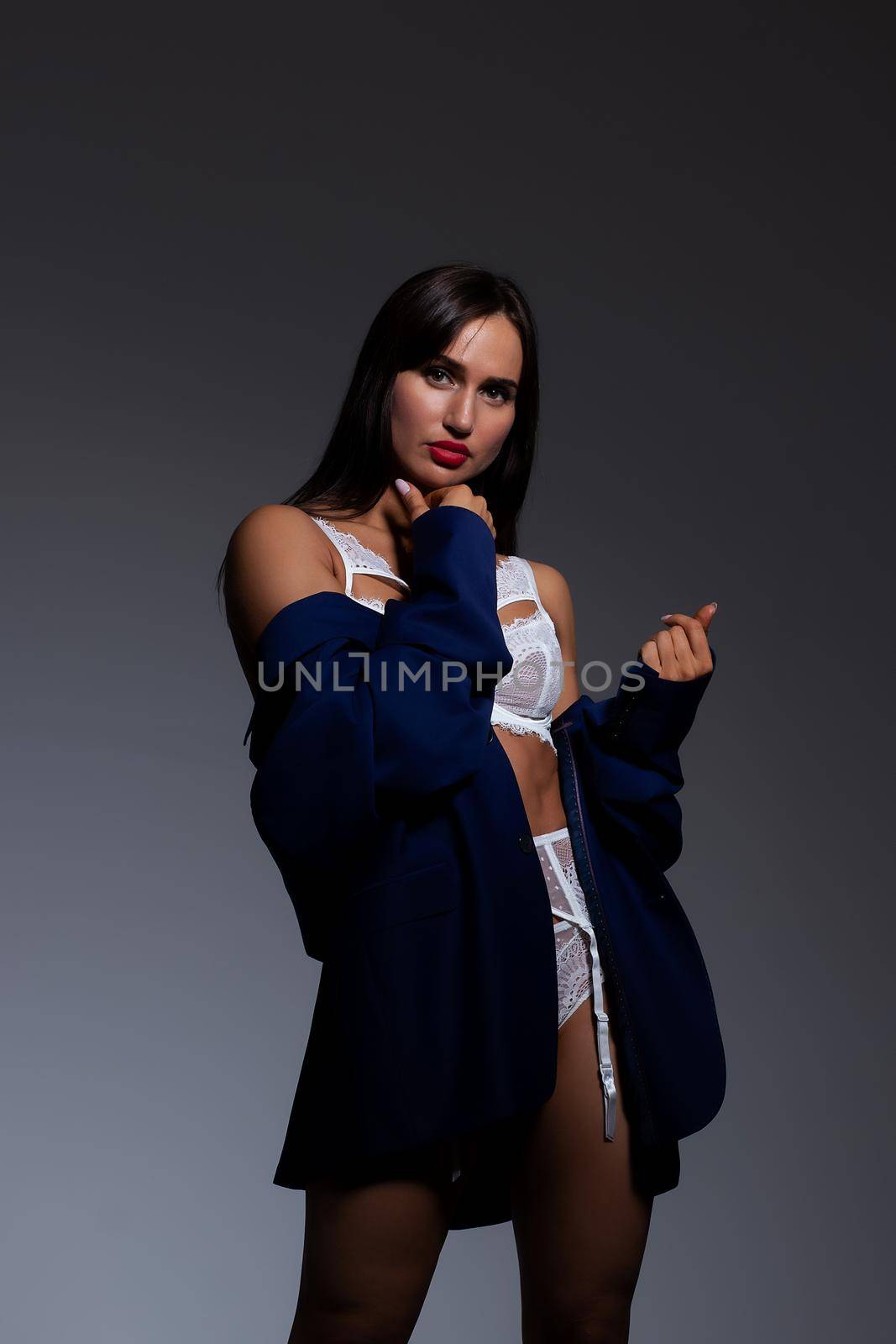 Back view alluring gorgeous brunette wearing white lace lingerie lowering jacket and looking at camera over shoulder during photo session in studio