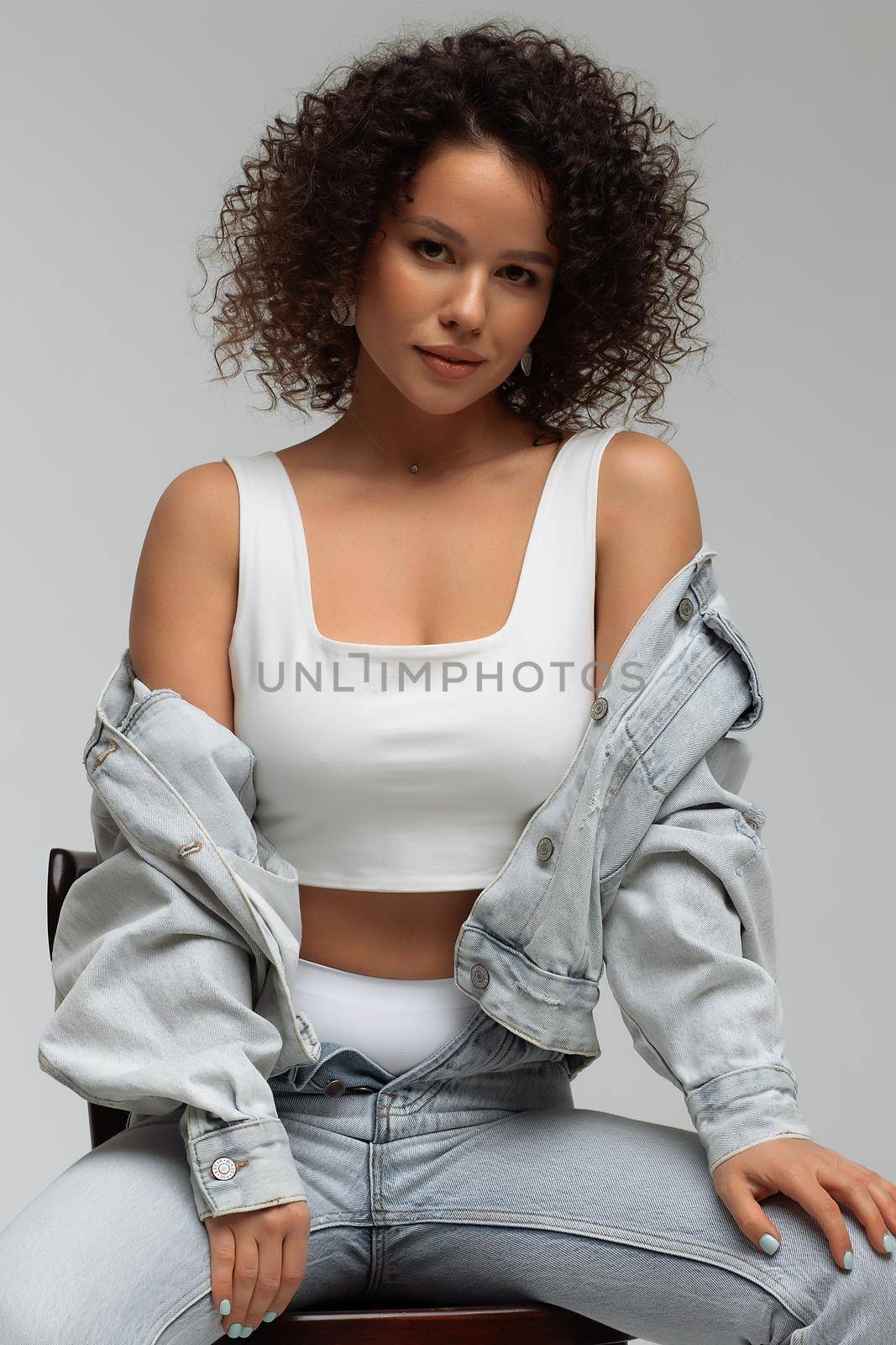 Full body of confident young barefoot female model with curly hairstyle wearing trendy denim jacket and jeans sitting on chair in white studio