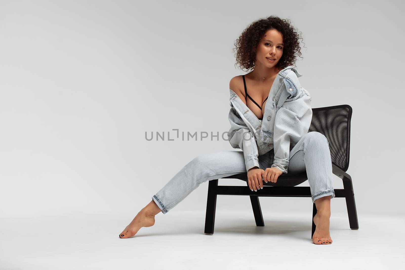 Full body of confident young barefoot female model with curly hairstyle wearing trendy denim jacket and jeans sitting on chair in white studio