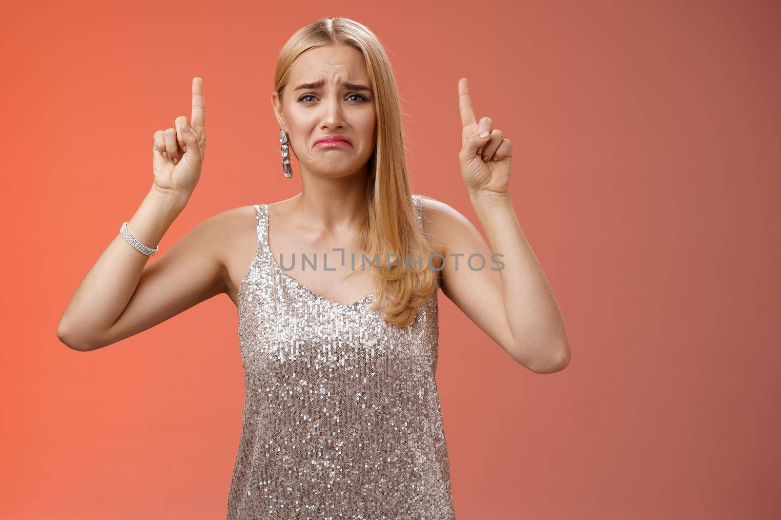 Upset whining immature spoiled blond rich girl in silver glittering dress pouting frowning gonna cry pointing up regret jealousy begging buy expensive shoes, standing red background displeased.