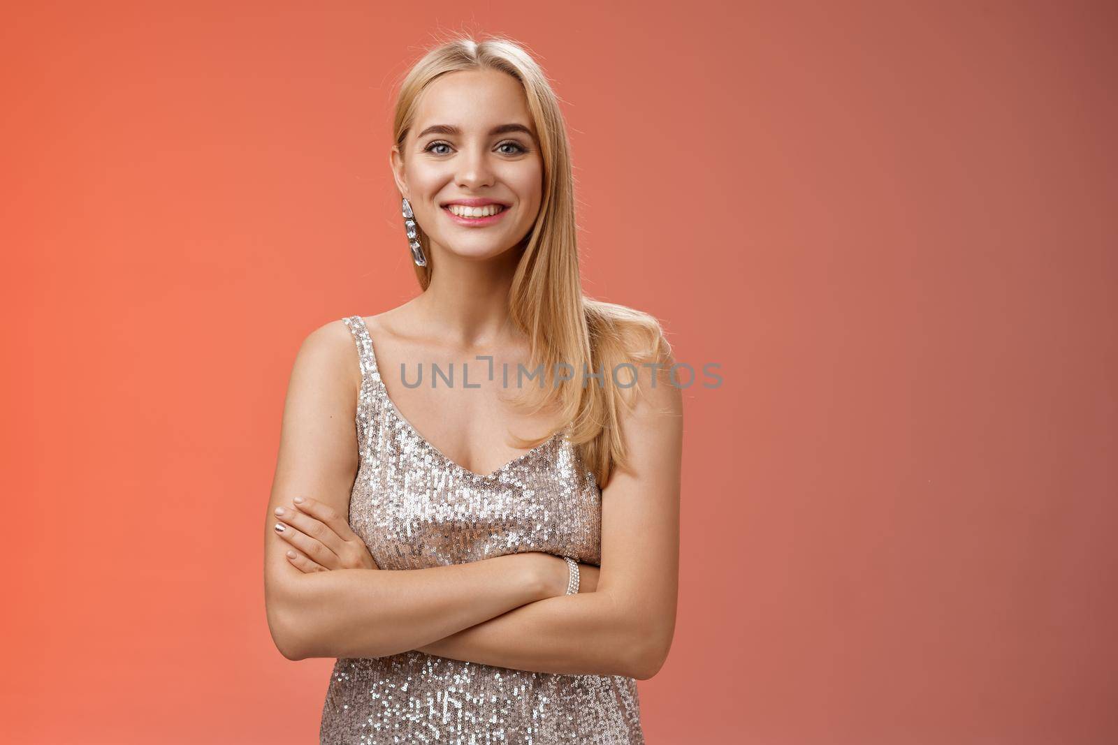Charming tender blond girlfriend in silver shiny elegant dress feel little cold standing prom night talking boyfriend dance floor hold hand crossed chest grinning having fun, standing red background.