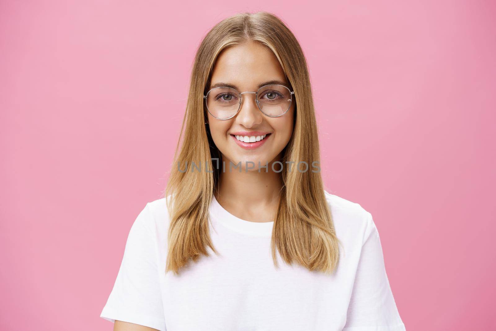 Creative and smart good-looking friendly female with tanned skin and little pimples wearing glasses and white t-shirt smiling broadly with perfect teeth standing plesant over pink wall. Copy space