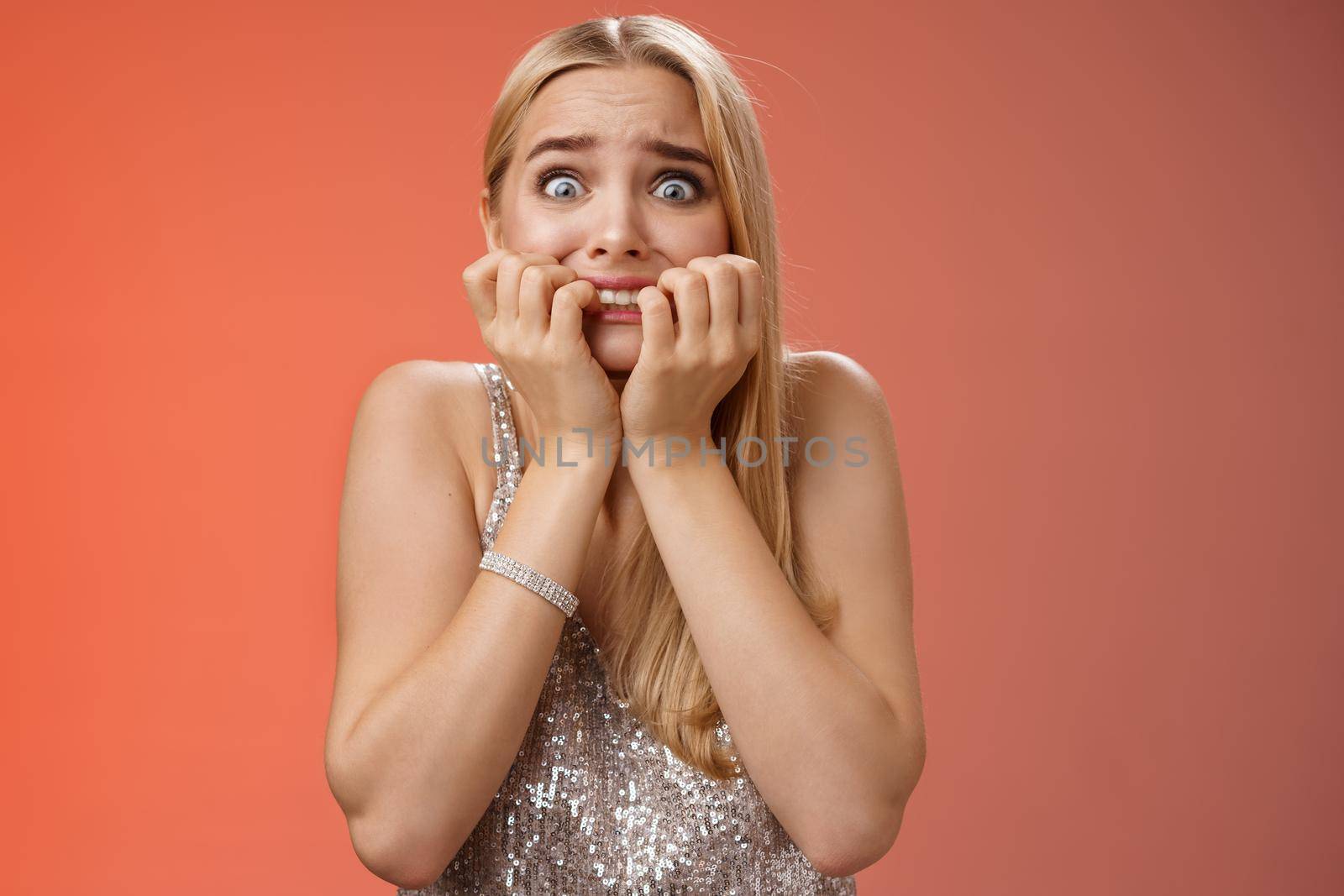 Frightened afraid panicking young cute blond woman biting nails pop eyes scared camera look terrified stunned speechless stare forward standing red background gasping trembling fear by Benzoix