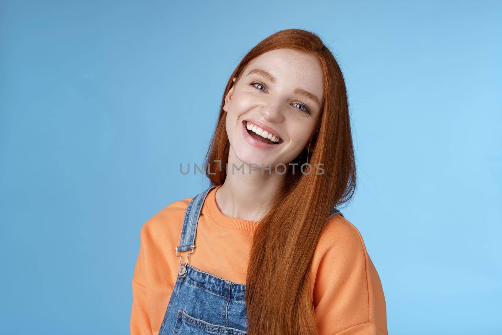 Positive outgoing lively redhead girl laughing joyfully having fun talking friendly friends tilting head chuckling joking funny life moments standing positive lucky blue background orange t-shirt. Lifestyle.