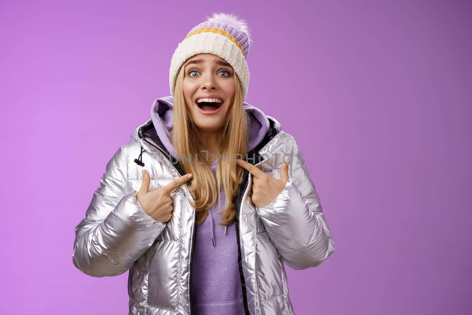 Surprised happy smiling joyful attractive woman pointing herself satisfied picked was chosen winner standing excited in silver shiny jacket winter hat cannot believe own luck, purple background.