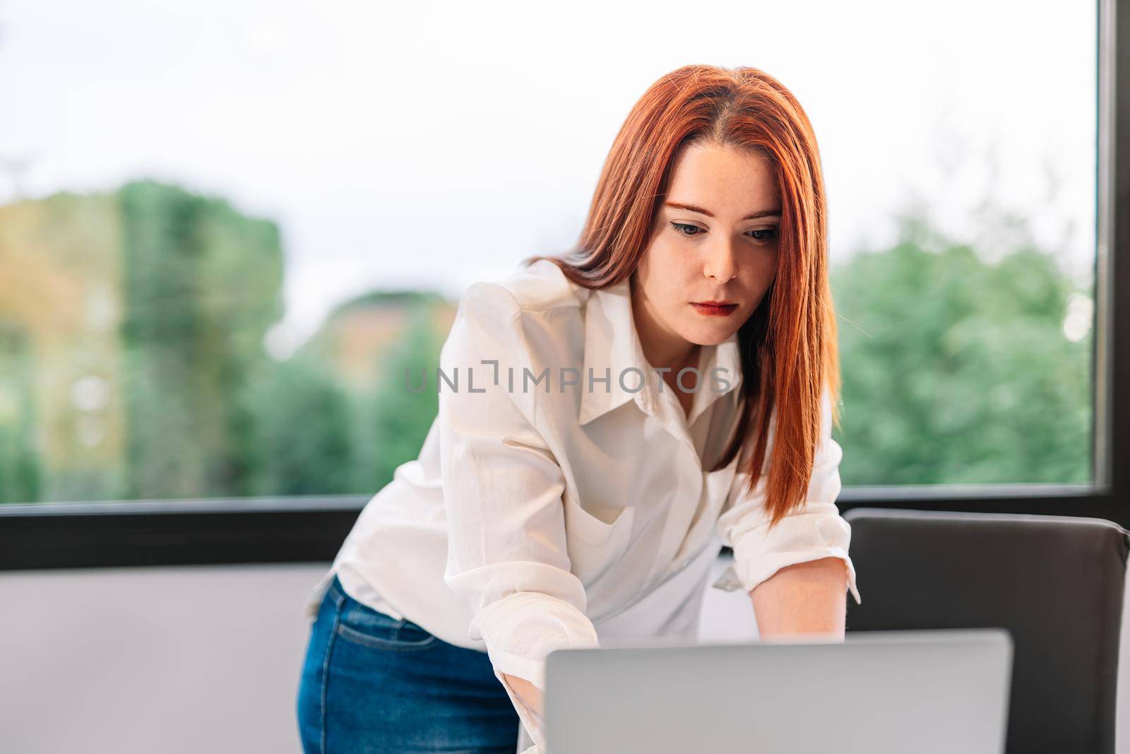 Attractive pretty young red-haired woman in white shirt and blue jeans standing looking at information on laptop while teleworking at home. Self-employed entrepreneur working from home on laptop. Large window in background with natural light.