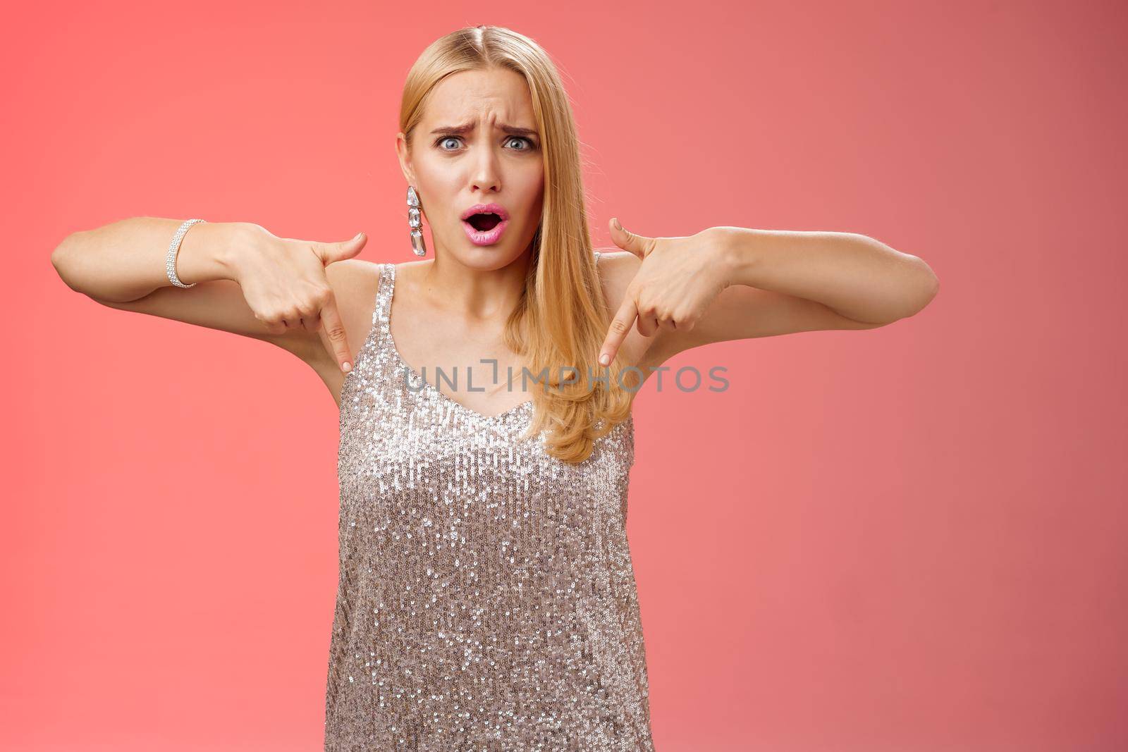 Upset disappointed fooled blond elegant girlfriend in silver dress frowning grimacing bothered pointing down displeased disliking shoes bought online store not match outfit, standing unhappy.