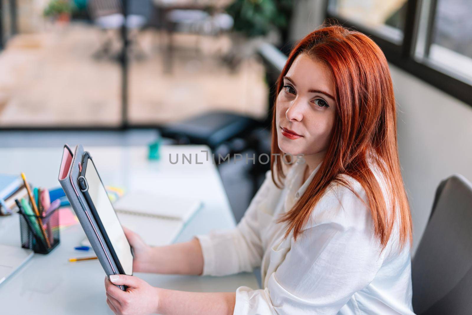 Portrait of a pretty young attractive girl with red hair in a white shirt using a tablet while teleworking at home. Enterprising young entrepreneur and self-employed girl working from home with tablet. White screen of tablet to paste any content. Large window in background with natural light.